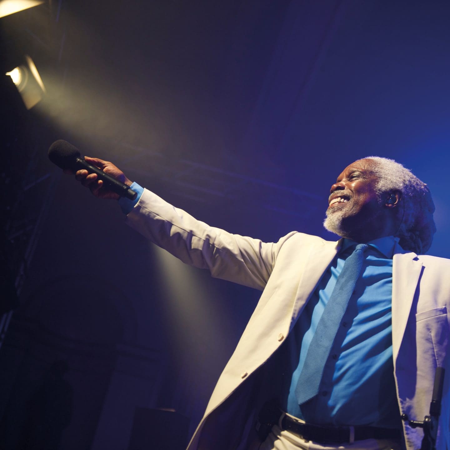 Picture of Billy Ocean (black male, with long grey hair in a pony tale and grey beard) smiling in a white suit with blue shirt and tie. Stood in stage lights holding a mike out to the audience. He has a glowing smile full of charisma.