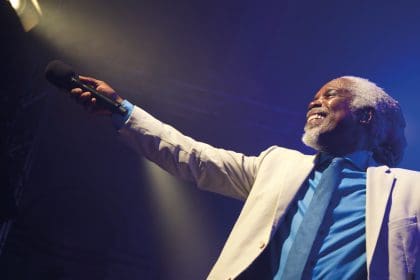 Picture of Billy Ocean (black male, with long grey hair in a pony tale and grey beard) smiling in a white suit with blue shirt and tie. Stood in stage lights holding a mike out to the audience. He has a glowing smile full of charisma.